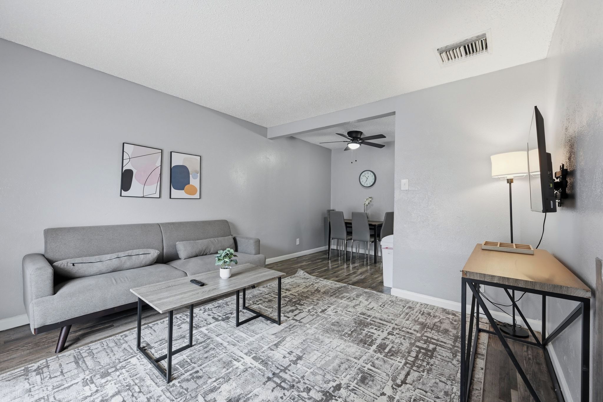 Una sala de estar moderna con un sofá gris, una mesa de centro, arte abstracto en la pared, un comedor con sillas, un ventilador de techo y un televisor montado.