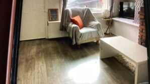 Ein Zimmer mit Holzboden, einem grauen Sofa mit orangefarbenem Kissen, einer weißen Bank und einem Fenster mit Blick ins Freie.