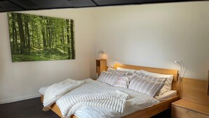 Un letto rifatto con cura, con una testata in legno, un comodino con una lampada e un grande quadro a tema foresta sulla parete.