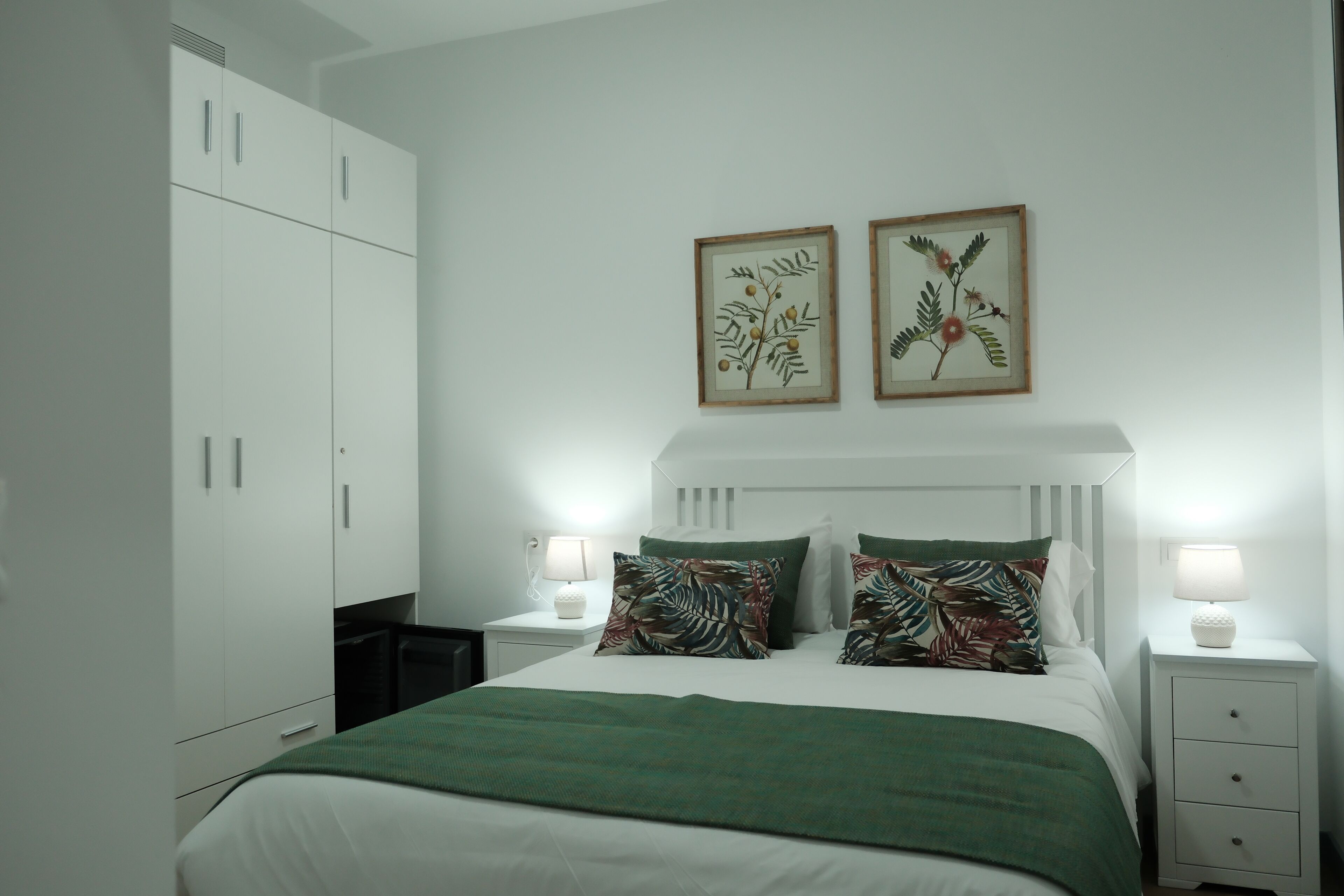 Un dormitorio con cama, mesitas de noche, un armario y dos láminas botánicas enmarcadas en la pared.