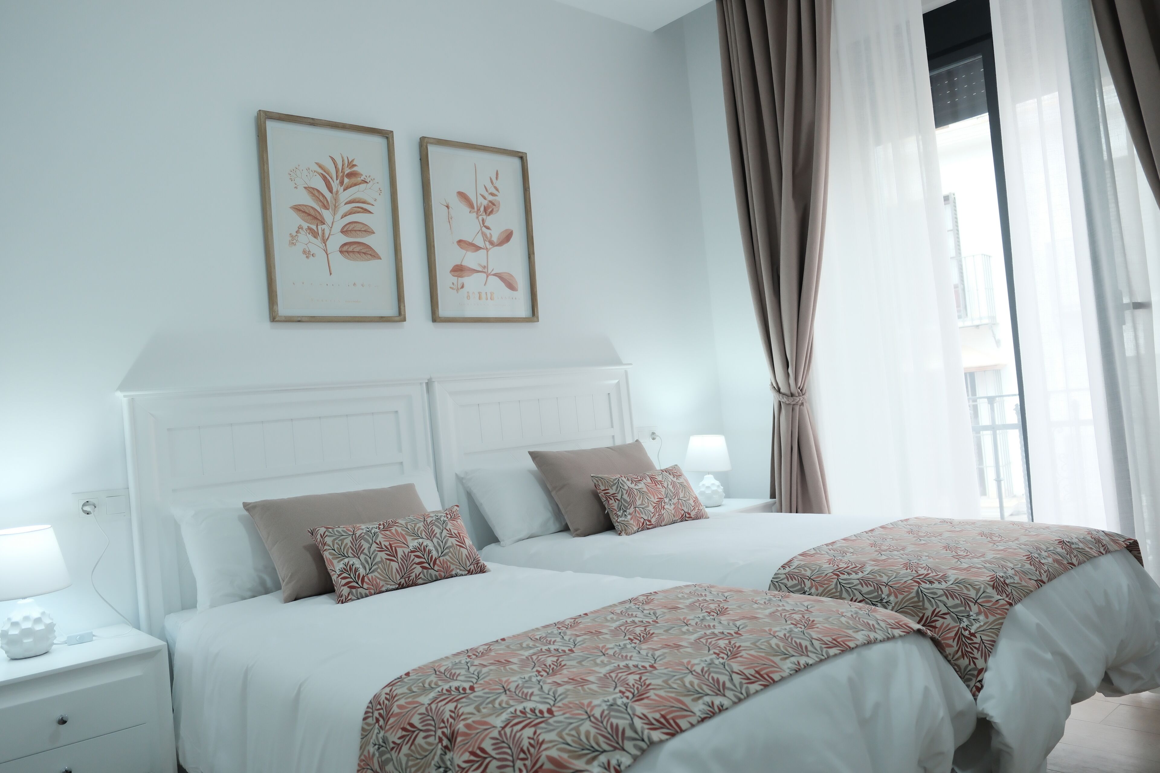 Una cama doble con cubrecama floral, cabecera blanca y dos impresiones botánicas enmarcadas en la pared.