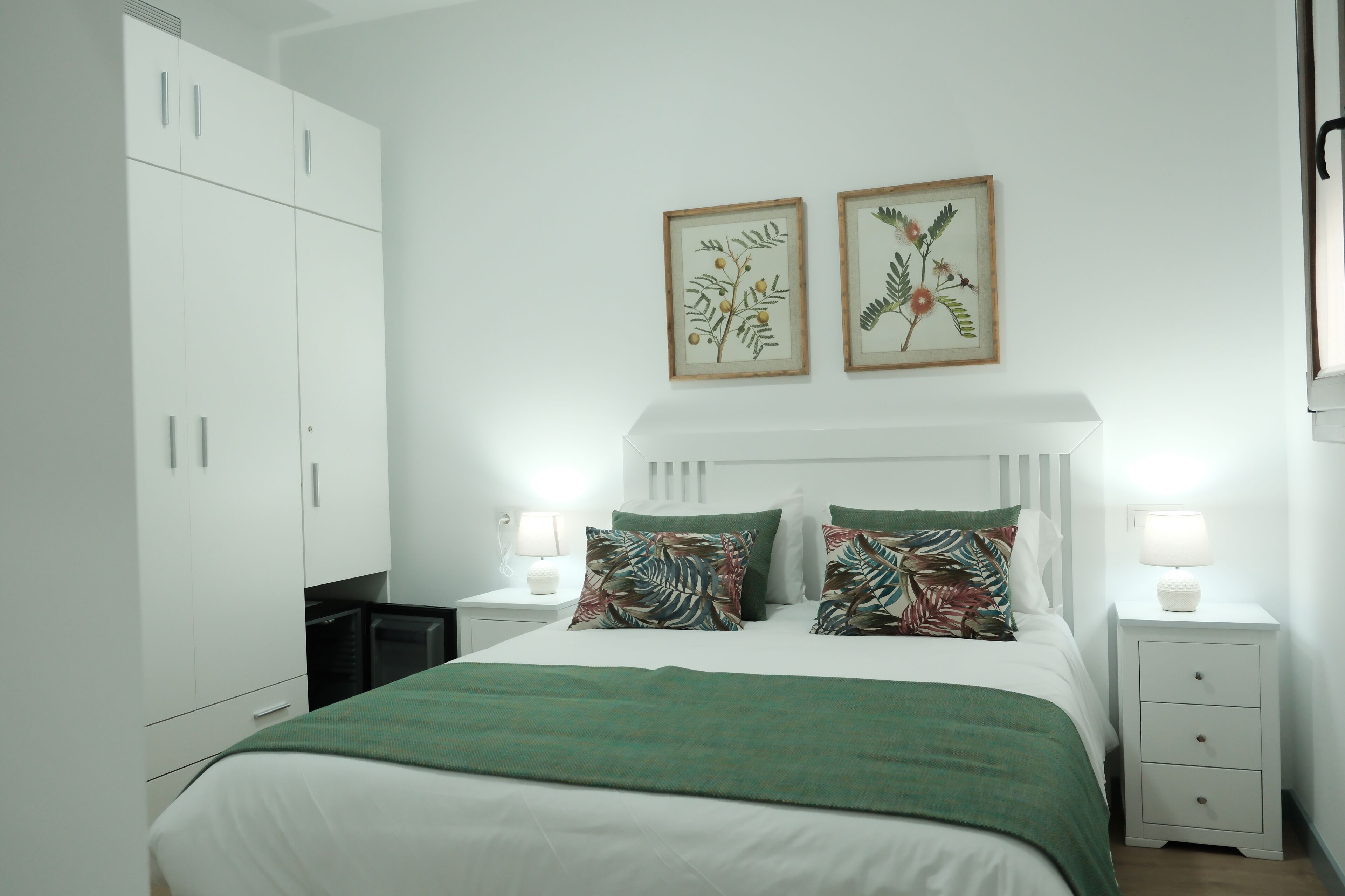 Een slaapkamer met een bed, een witte kledingkast, twee nachtkastjes met lampen en lijstwerken met plantenprints aan de muur.