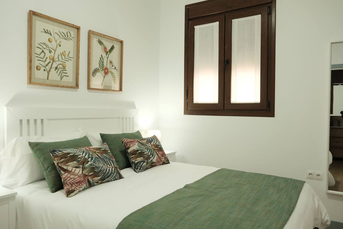A bed with a green bedspread, white pillows, and two framed botanical prints on the wall.