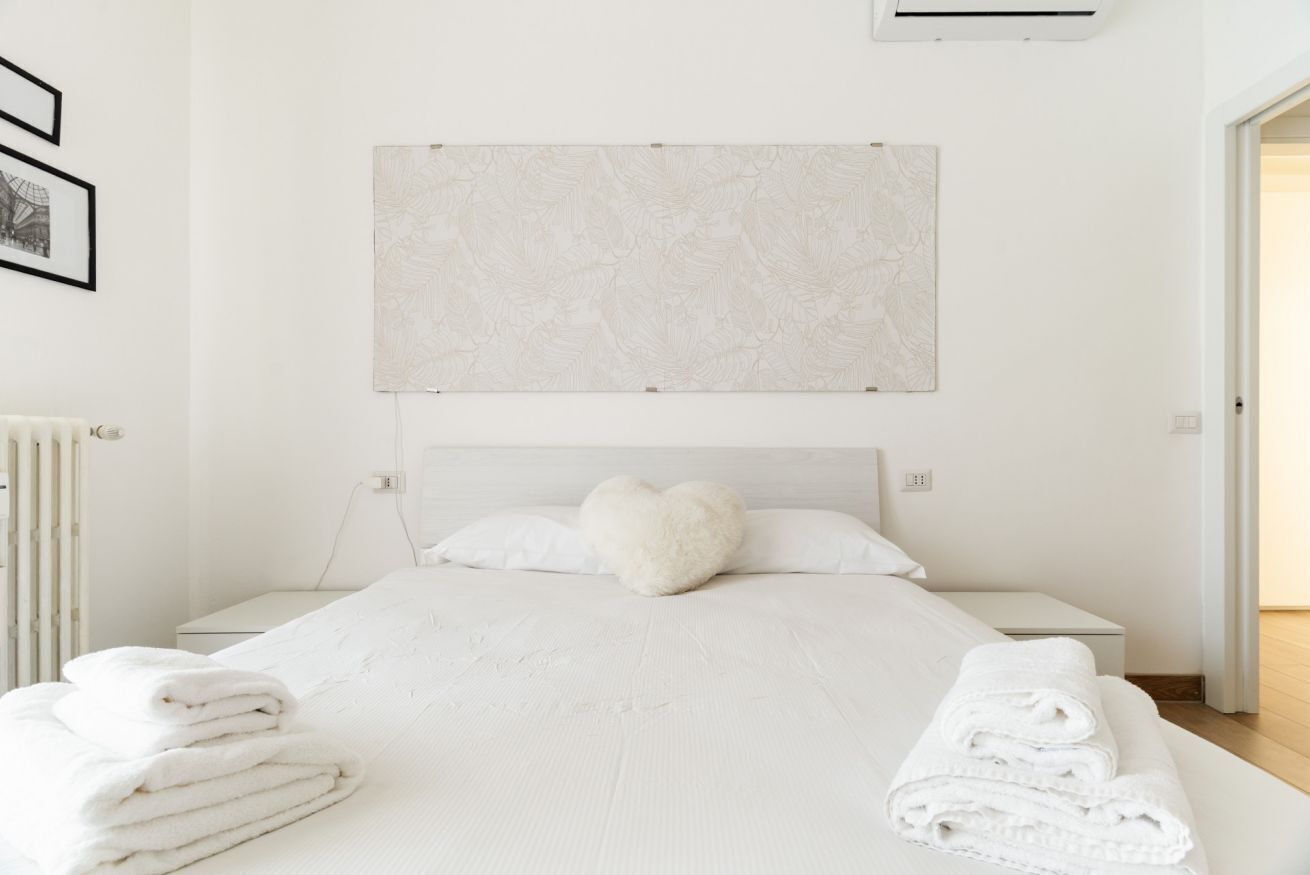 A bedroom with a bed, white linens, a heart-shaped pillow, a wall-mounted artwork, and a radiator.