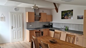Een moderne keuken met houten kastjes, een eettafel en stoelen. Er hangt een klok aan de muur en een schilderij boven de gootsteen.