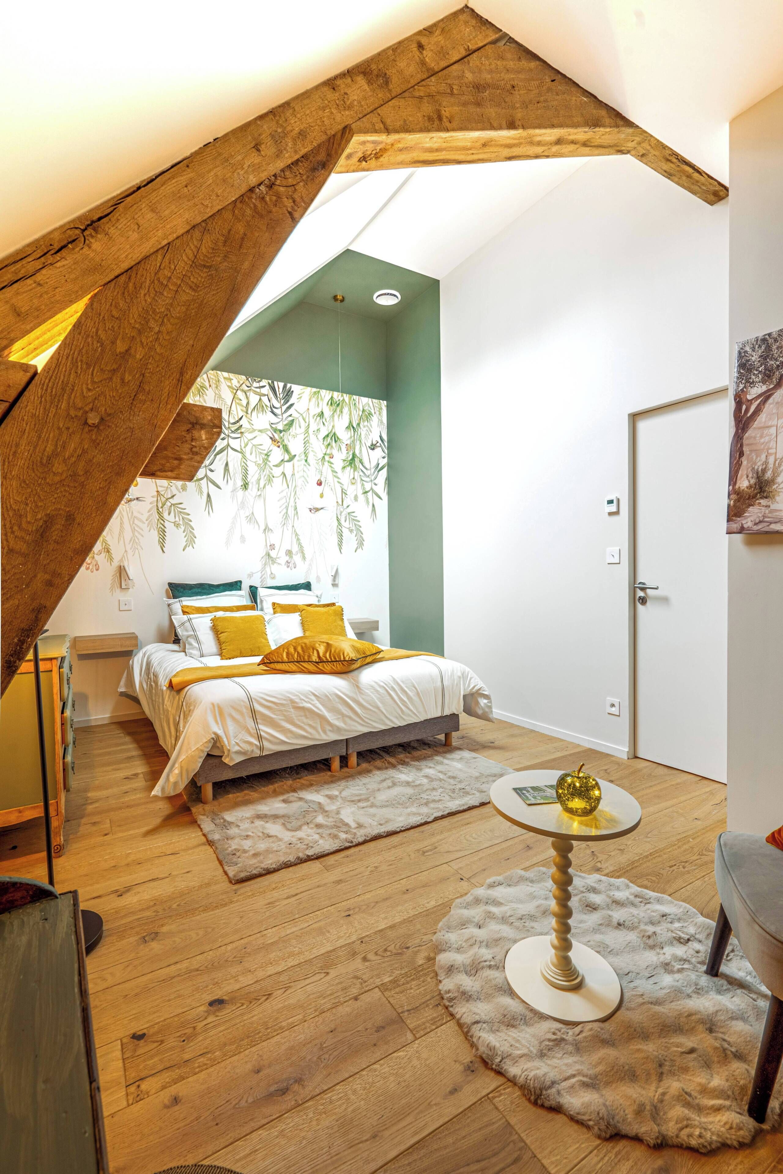 Quarto com teto inclinado, cama com almofadas amarelas, mesa redonda com um globo e porta com um quadro.