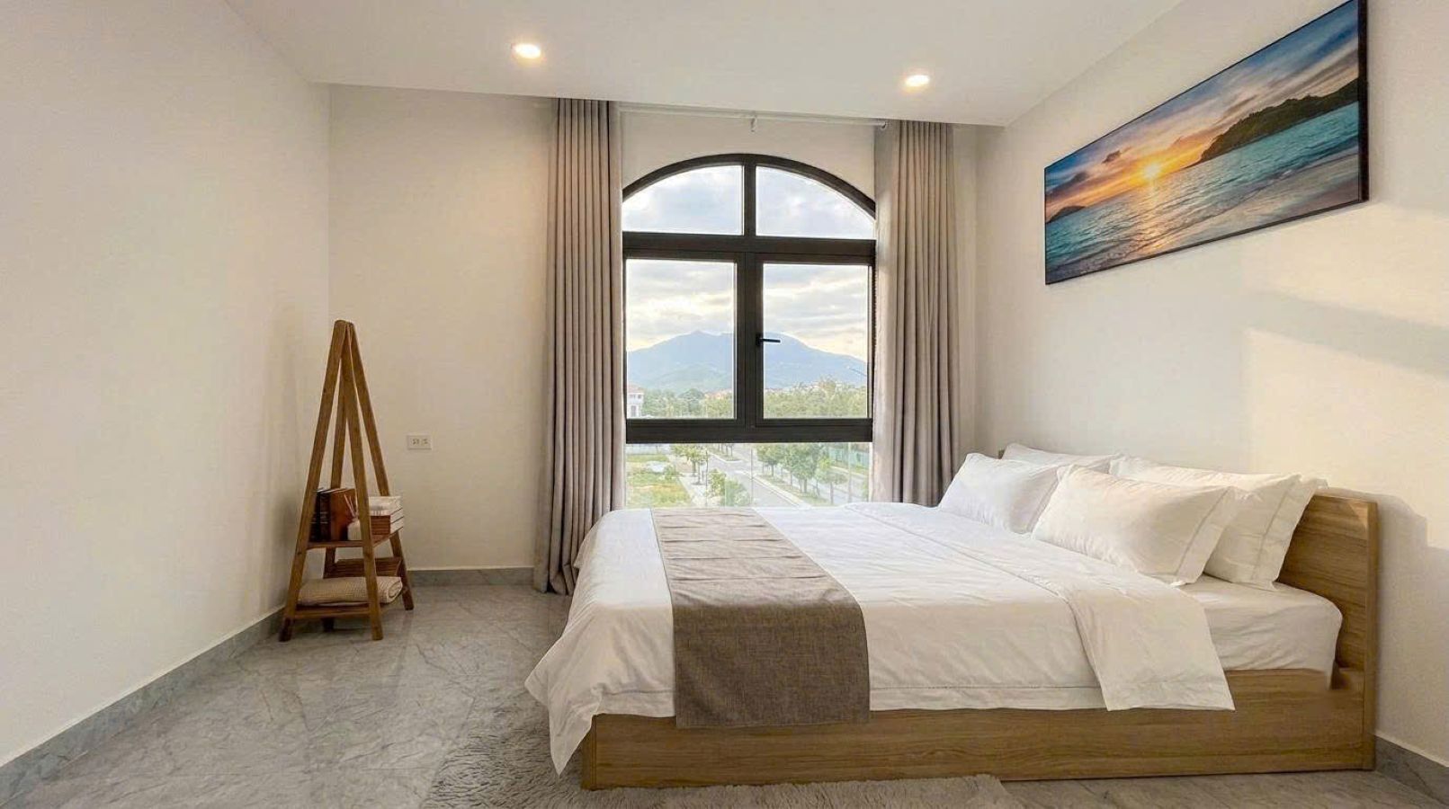 A hotel room with a bed, a window, a painting, and a wooden ladder.