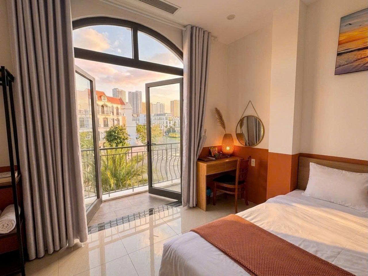 A hotel room with a bed, a desk, a chair, a window with curtains, and a view of the cityscape.