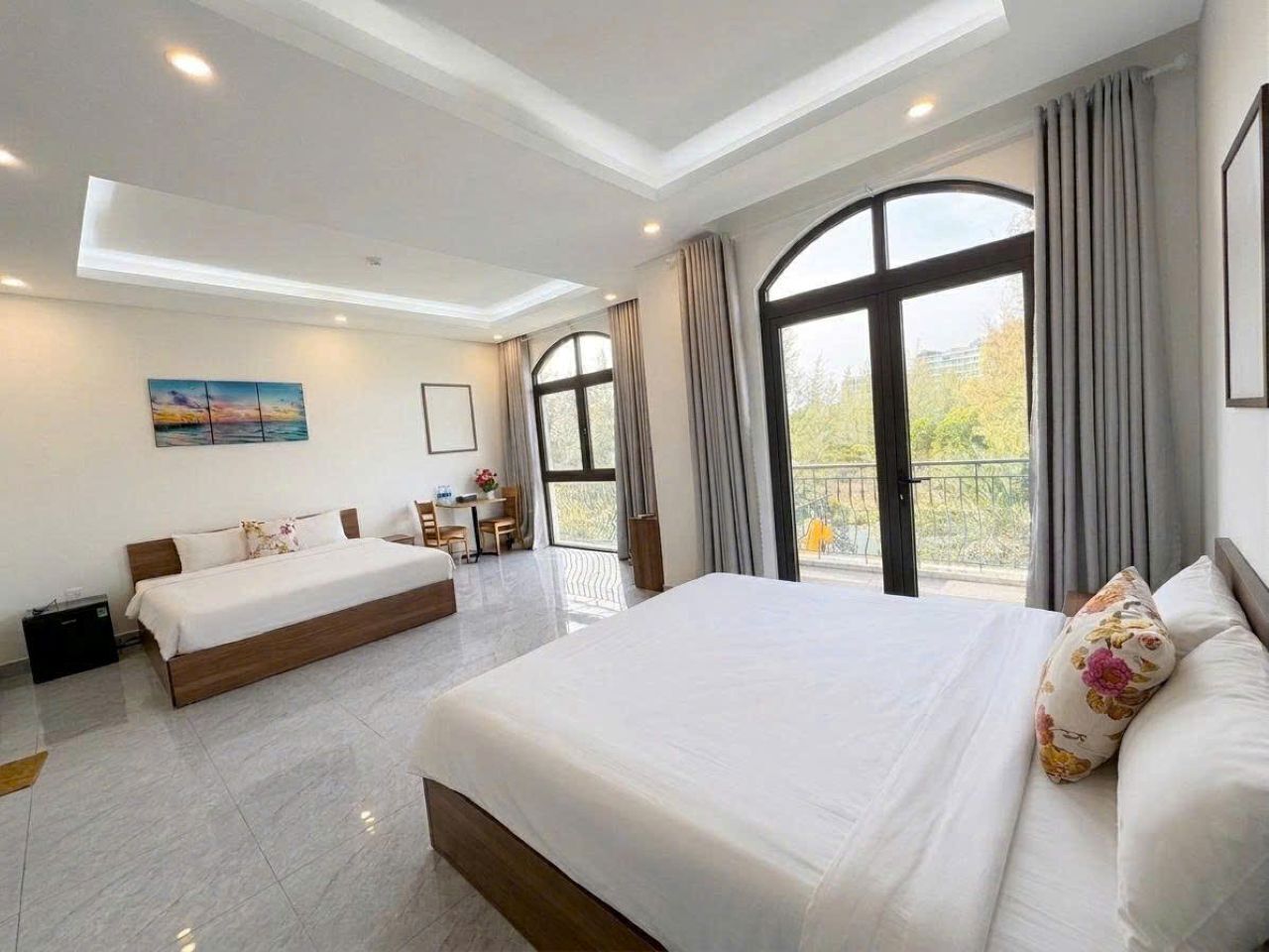 A hotel room with a large bed, a smaller bed, a desk, and a balcony with curtains.
