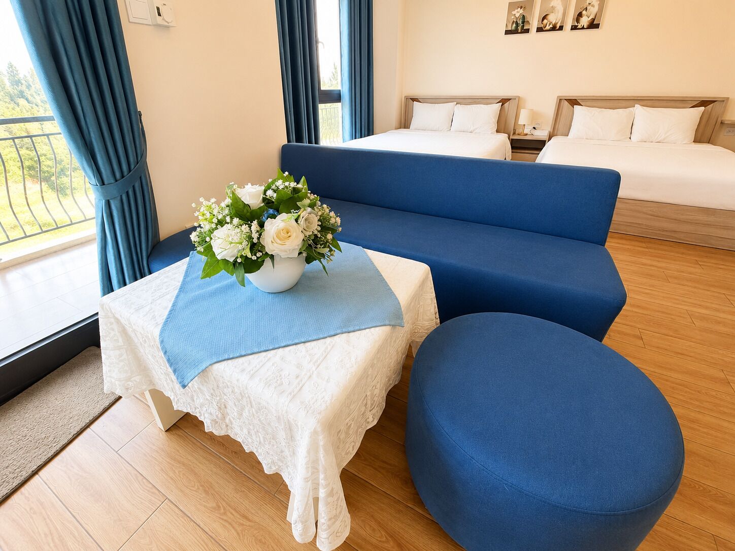 Habitación de hotel con un sofá azul, un otoman redondo, una mesita con mantel de encaje blanco y un jarrón con flores.