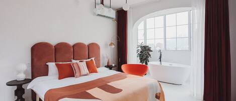 A modern bedroom with a large bed, a side table, a chair, a bathtub, and a window with curtains.