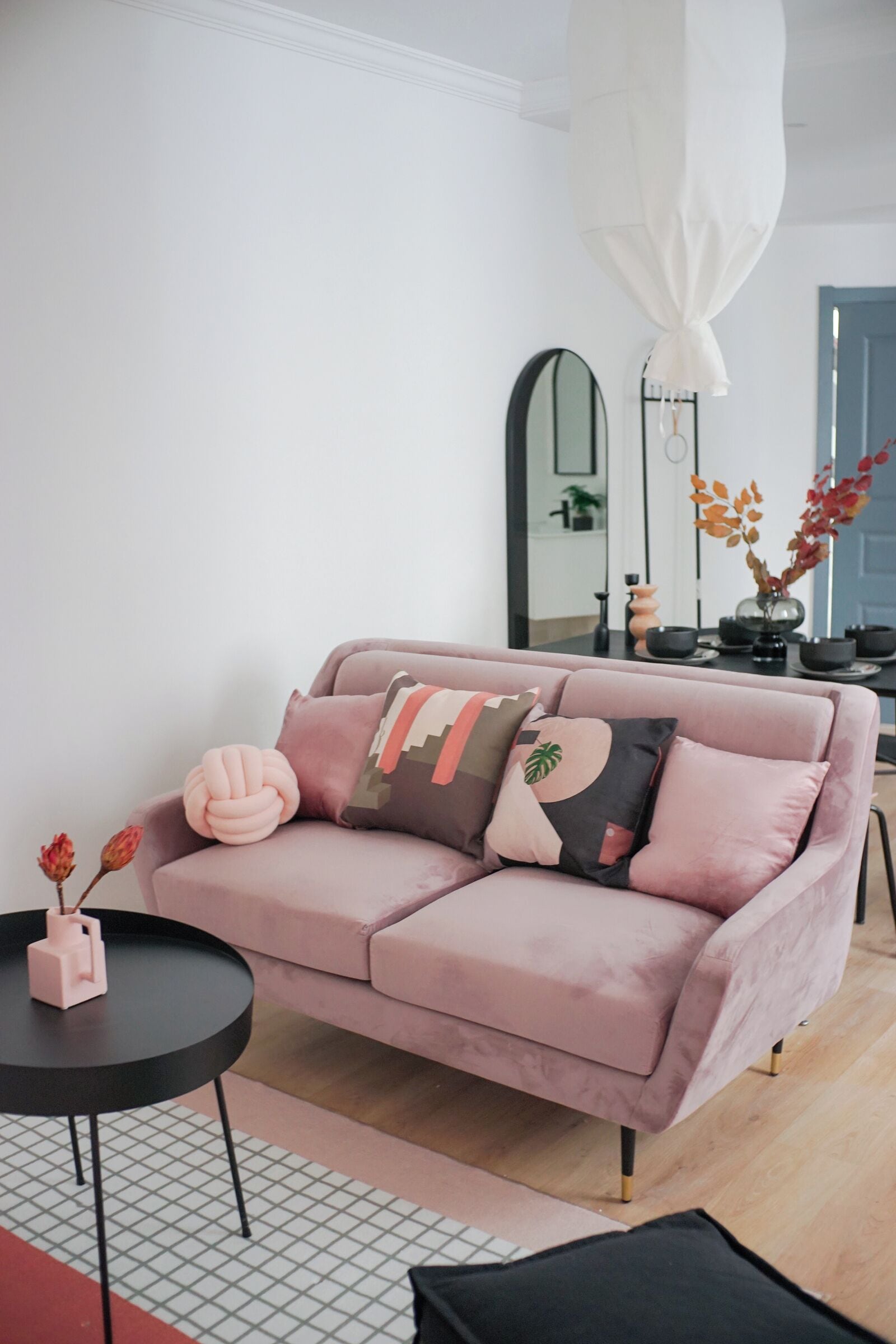 Un canapé rose agrémenté de coussins décoratifs, une petite table noire sur laquelle est posé un vase, et un miroir accroché au mur.