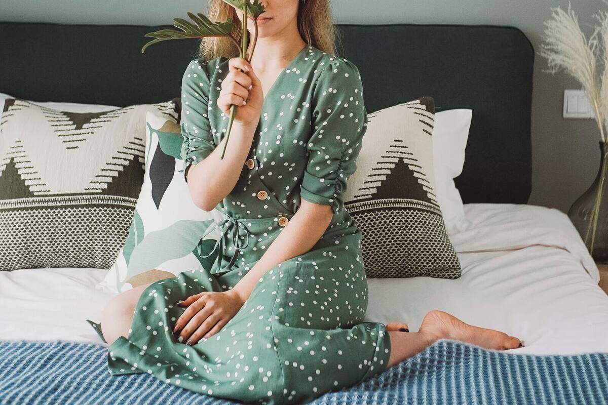Una persona sentada en una cama con un cojín estampado, un cuadro en la pared y un florero con plantas.