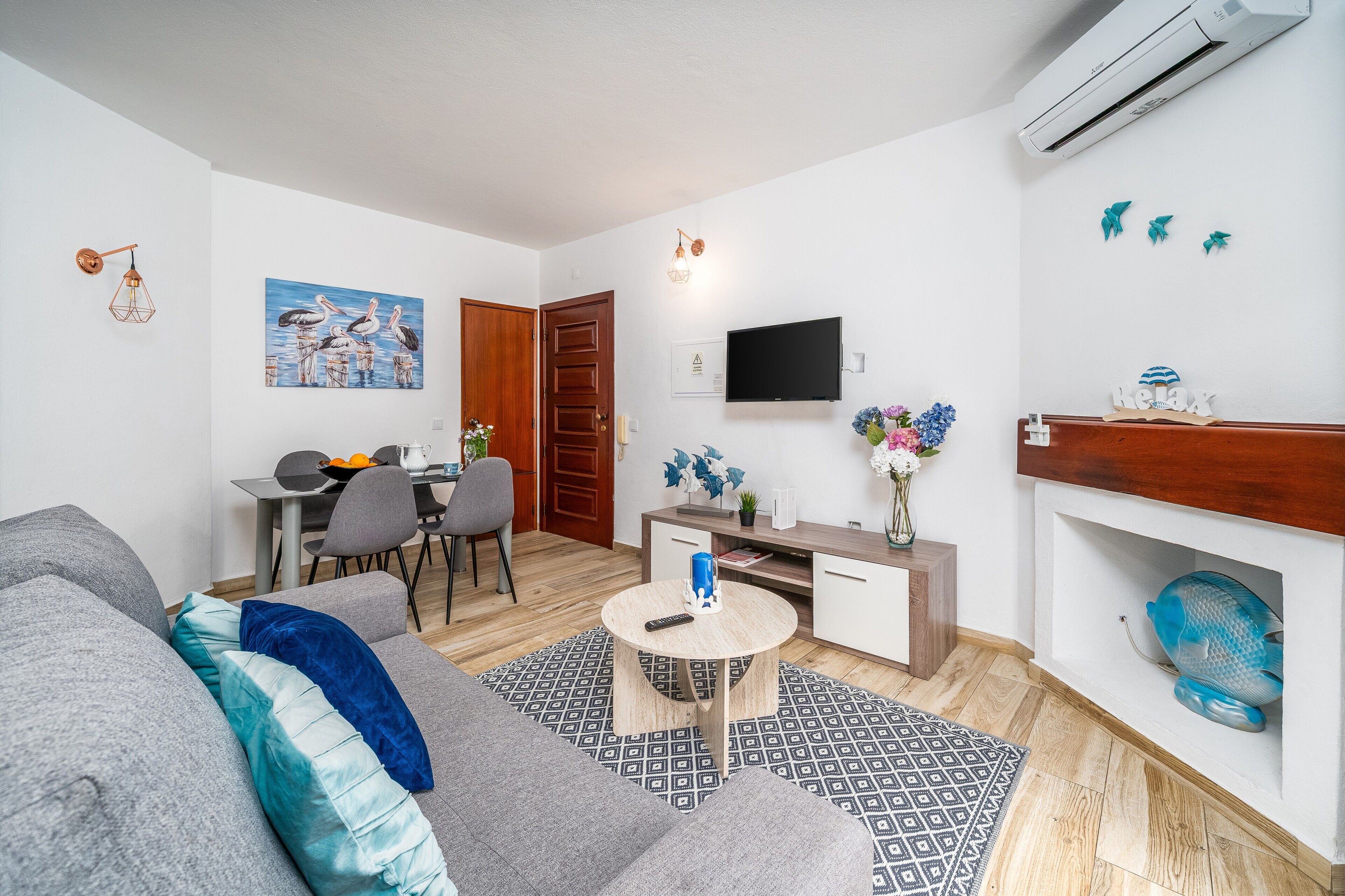 Una sala de estar con un sofá gris, una mesa redonda, un televisor, una chimenea y un comedor con una mesa y sillas.