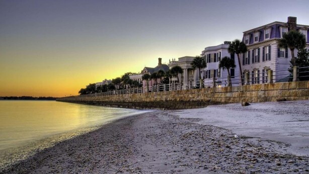 Una propiedad frente a la playa con un muro de piedra, una hilera de palmeras y un edificio de estilo arquitectónico clásico.
