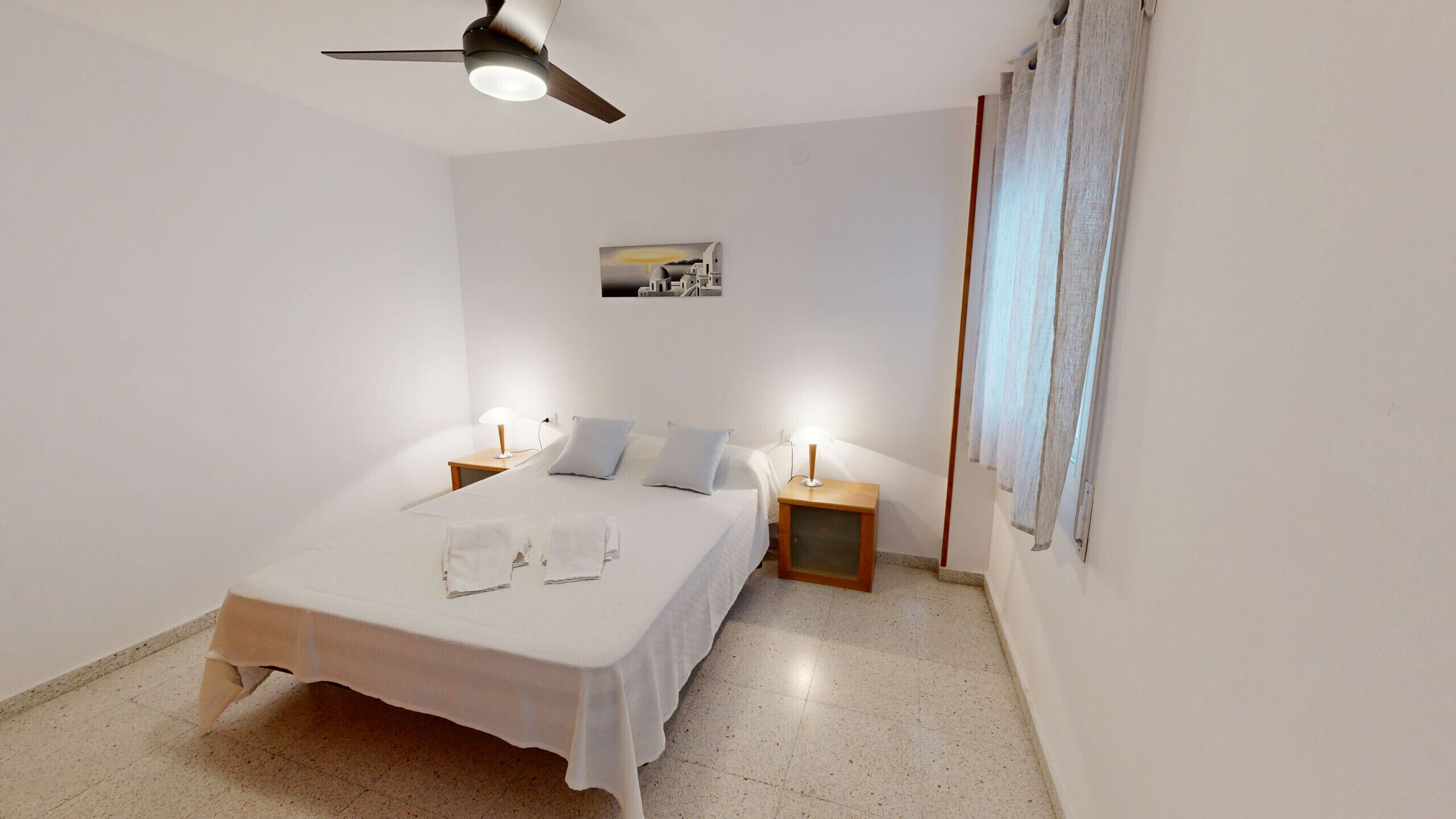 A bedroom with a bed, two bedside tables with lamps, a ceiling fan, and a painting on the wall.