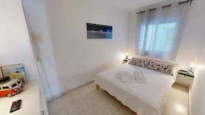 Een slaapkamer met een bed, kussens, een bedlampje, een wandklok, een ingelijste foto en een klein tafeltje met een lamp.