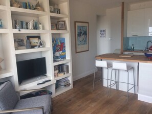 Een moderne woonkamer met een televisie, boekenkast en een kleine keuken met barstoelen.