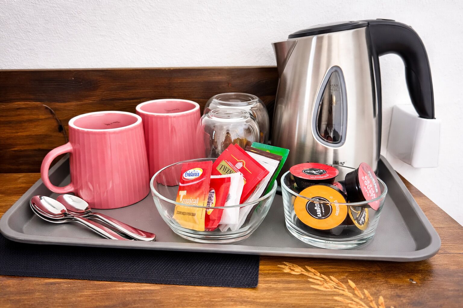 A tray with a kettle, pink mugs, a glass bowl with coffee packets, and a glass cup with a lid.