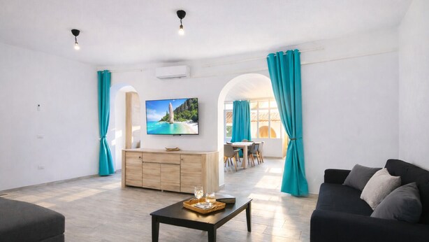 A spacious living room with a flat-screen TV, a wooden cabinet, a dining area with a table and chairs, and a black sofa. - Bonòra suites (Tortolì)