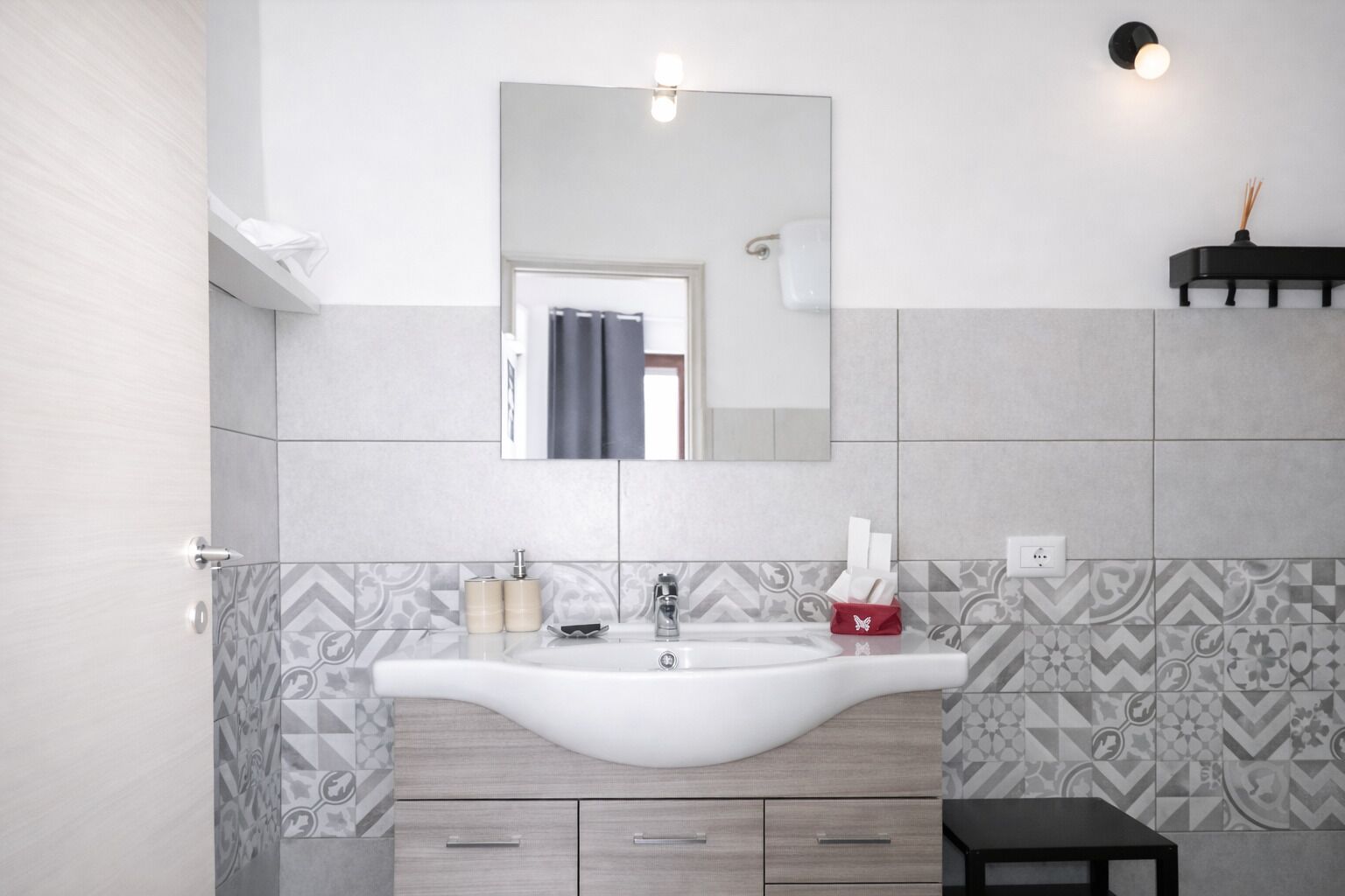 A bathroom with a white sink, a mirror above it, patterned tiles, a small stool, and a shelf with a candle.