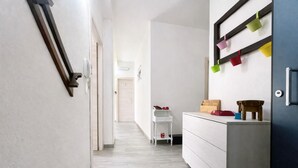 A hallway with a white wall, a wooden floor, a white side table with a red bag, a wooden chair, and a blue stool. - Bonòra suites (Tortolì)