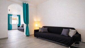 A modern living room with a black sofa, teal curtains, a glass coffee table, and a lamp. - Bonòra suites (Tortolì)