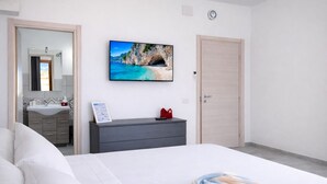 A hotel room with a bed, a dresser, a TV on the wall, a bathroom with a sink and mirror, and a door. - Bonòra suites (Tortolì)