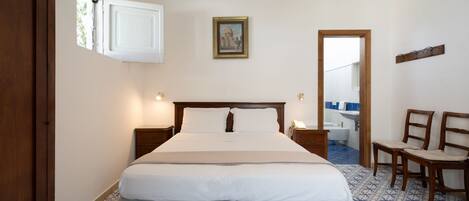 A bedroom with a bed, bedside tables, a mirror, a picture on the wall, and a bathroom visible through an open door.