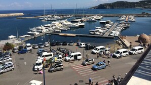 A marina with boats docked, a parking area with cars and vans, and a coastal landscape in the background.