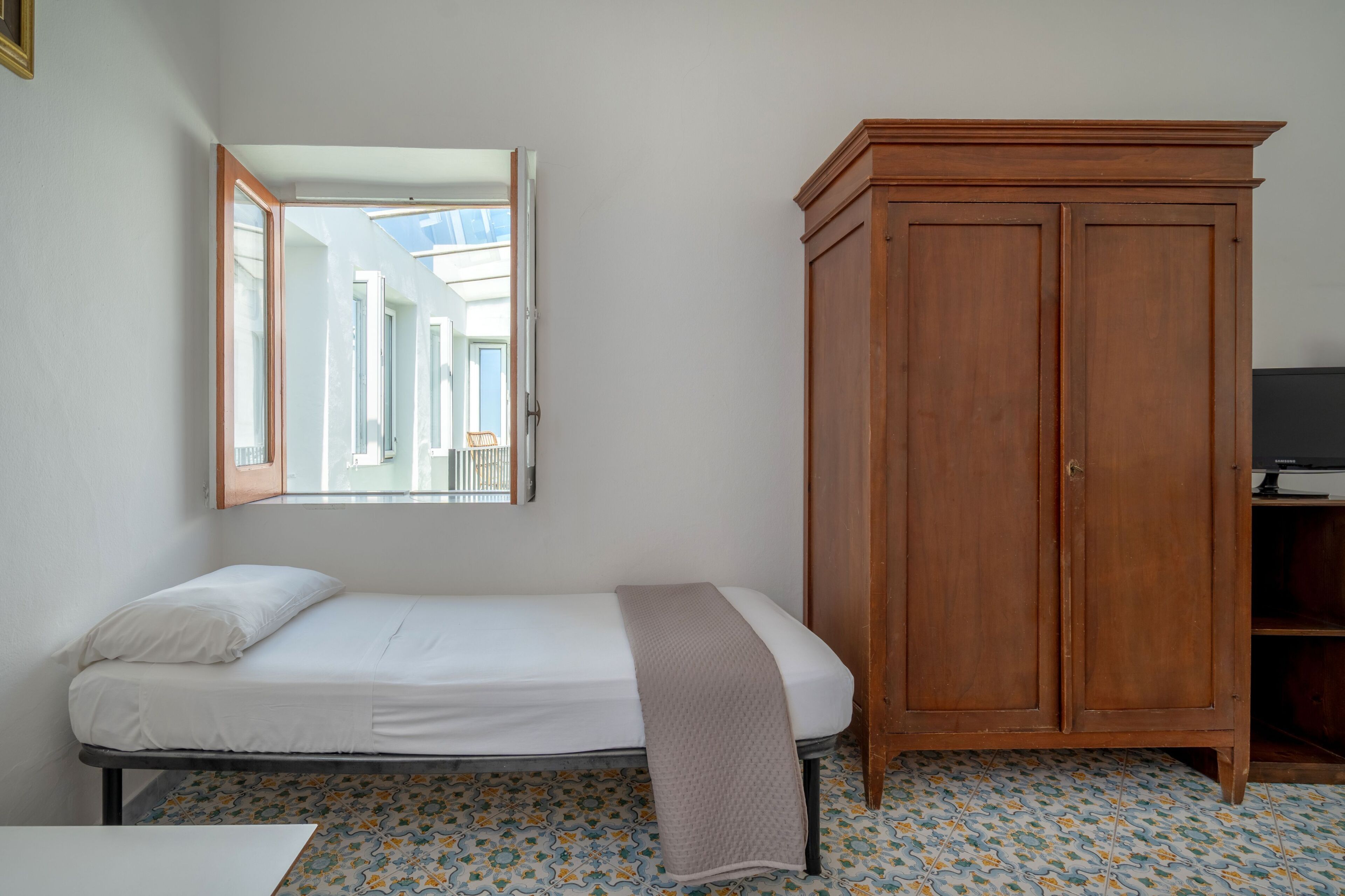 Een kamer met een enkel bed, een kledingkast, een spiegel en een betegelde vloer.