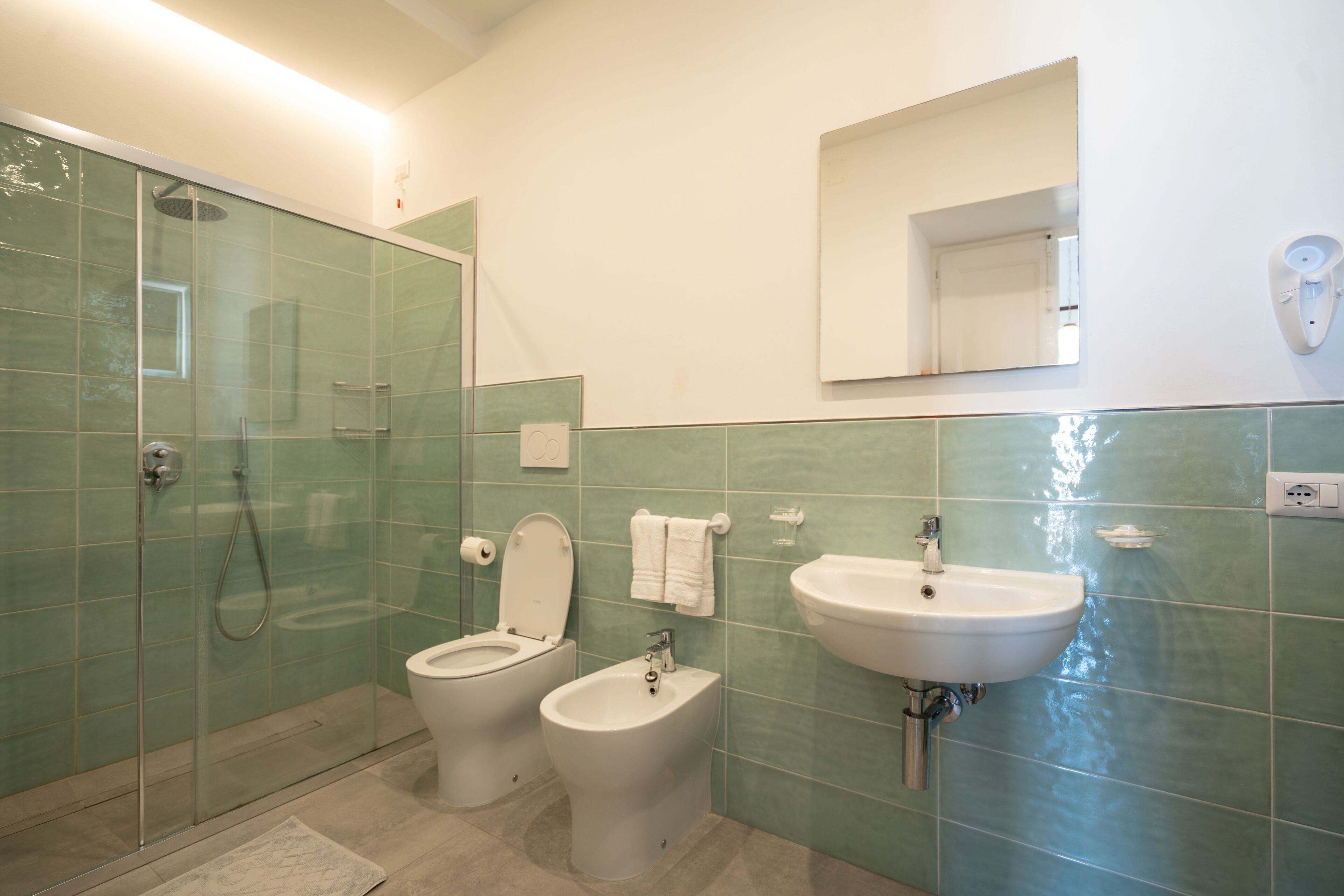 A bathroom with a glass-enclosed shower, two toilets, a bidet, a sink, and a mirror.