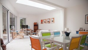 A bright, modern living room with a glass dining table, colorful chairs, a sofa, and a TV.