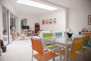 A bright, modern living room with a glass dining table, colorful chairs, a sofa, and a TV.