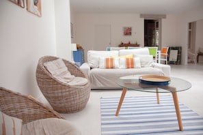 Un salon avec un fauteuil en osier, une table avec un plateau en verre, un tapis à rayures et un canapé blanc agrémenté de coussins.