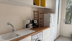 A compact kitchen with a sink, coffee maker, blender, wooden shelf, and a washing machine.
