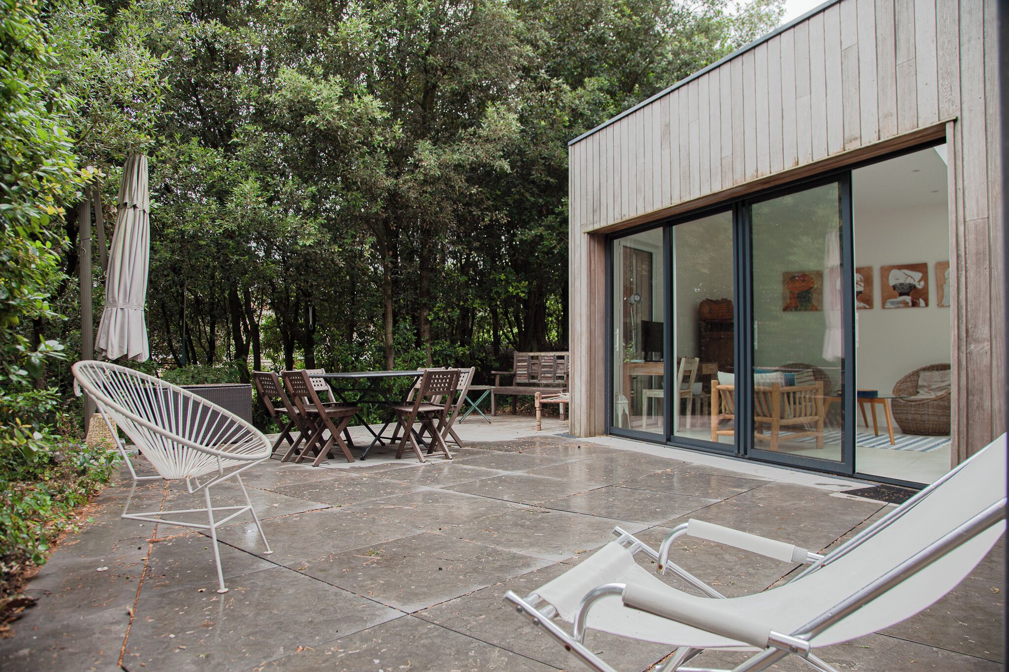 Un patio al aire libre con una mesa, sillas y una hamaca colgante, con un ventanal de vidrio que da a un espacio interior.