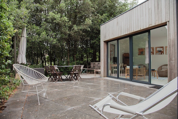Une terrasse extérieure avec une table et des chaises, un fauteuil suspendu et une porte vitrée donnant sur un espace intérieur.