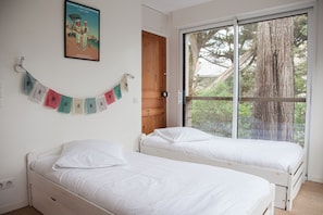 Une chambre avec deux lits simples, une porte en bois, une fenêtre donnant sur des arbres et une affiche accrochée au mur.