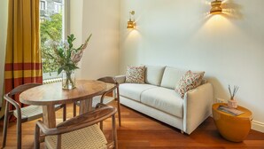 Ein Zimmer mit einer Couch, einem runden Holztisch und Stühlen. Es gibt Wandleuchten und ein Fenster mit Vorhängen.
