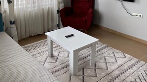A living room with a white table, a red chair, a TV mounted on the wall, and a painting above the TV. - kasapoglu ceren pansiyon  (Bursa)