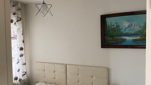 A bedroom with a bed, a ceiling fan, a painting, and a small wooden side table. - kasapoglu ceren pansiyon  (Bursa)