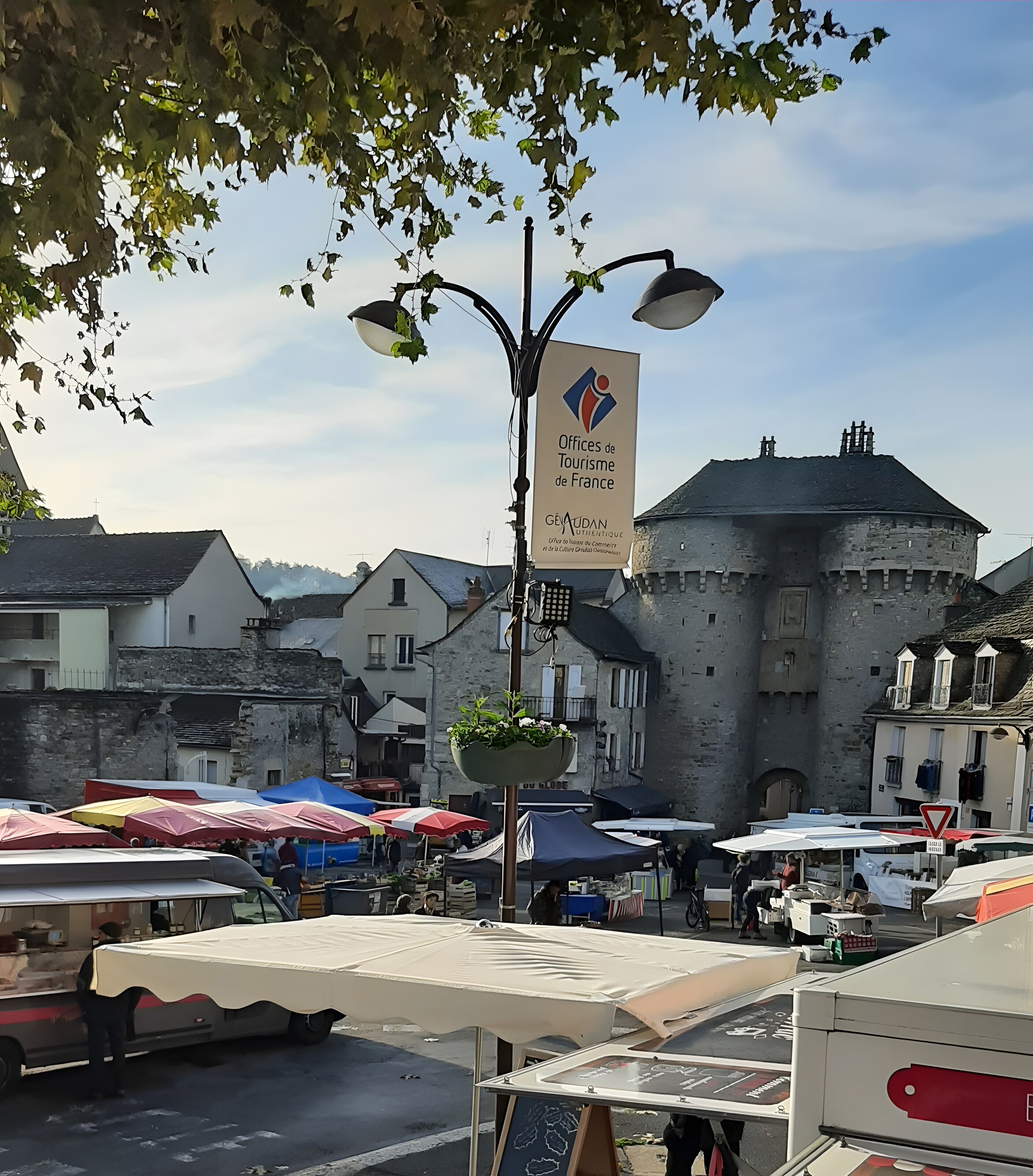 A medieval-style building with a stone facade, a street market with stalls, and a sign for 'Offices de Tourisme de France'.