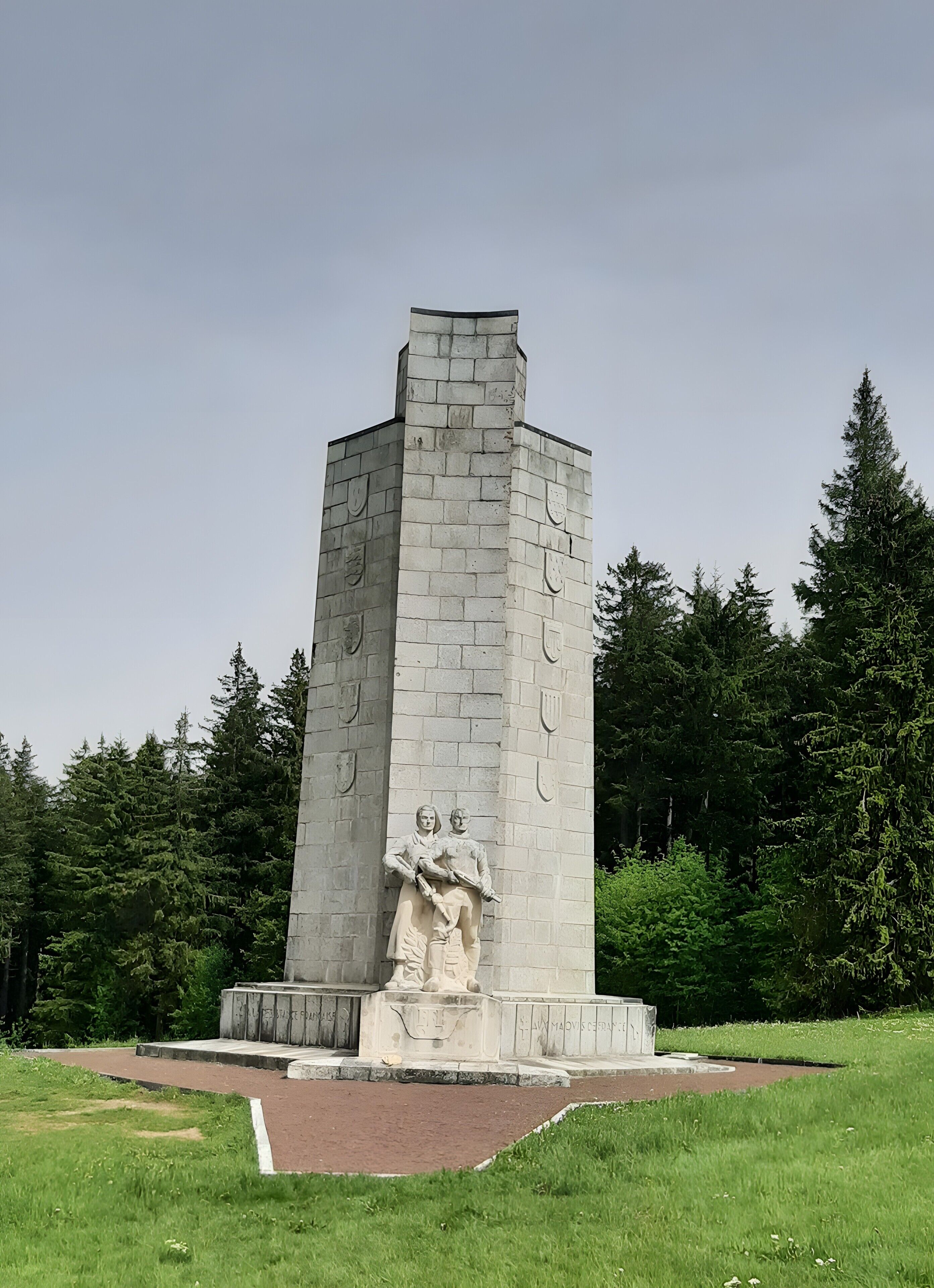 Ein hohes, rechteckiges Steinmonument mit einem zentralen Reliefbild von zwei Figuren, umgeben von befestigtem Untergrund und Grünpflanzen.