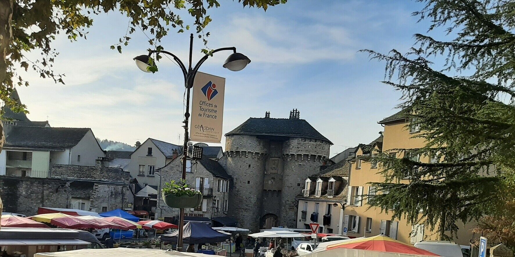 En historisk byplads med et markant stenhus, markedsboder og et skilt for Offices de Tourisme de France.