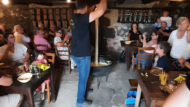 Un coin repas rustique avec des tables et des chaises en bois, un grand pot noir posé sur le sol et des personnes assises autour des tables.