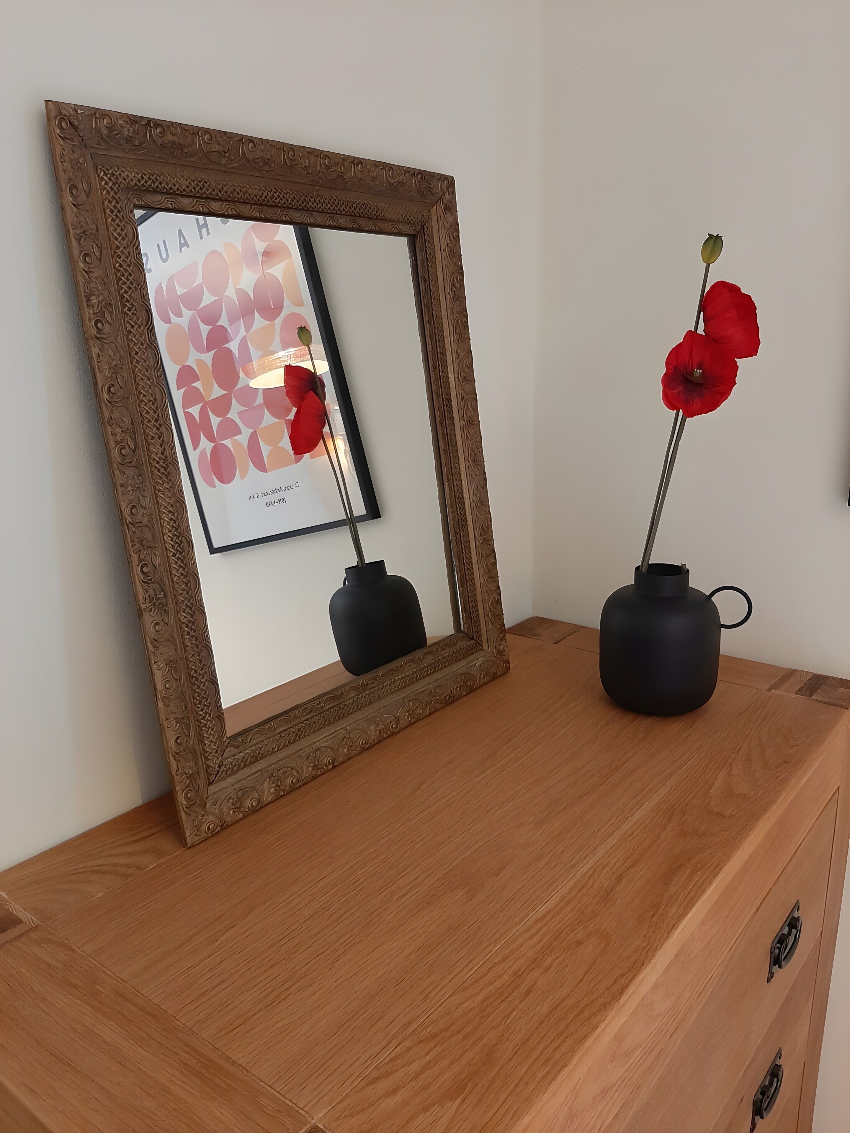 A mirror with an ornate frame, a framed artwork, and two black vases with red flowers on a wooden dresser.