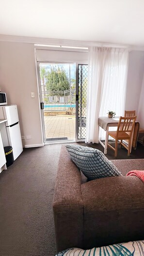 A room with a sofa, a dining table with chairs, a small kitchen area with a fridge, and a sliding glass door leading to an outdoor area with a pool. - Pool View Studio | 3 Min Walk to Beach | Marina & Foreshore (Mandurah)