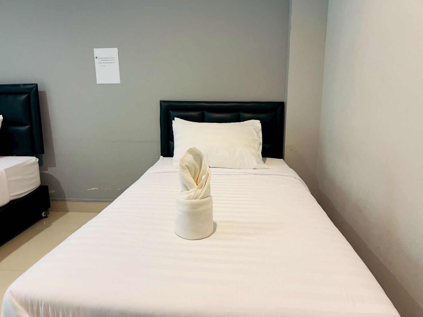 A single bed with a neatly folded towel, a dark headboard, and a plain wall with a small sign.