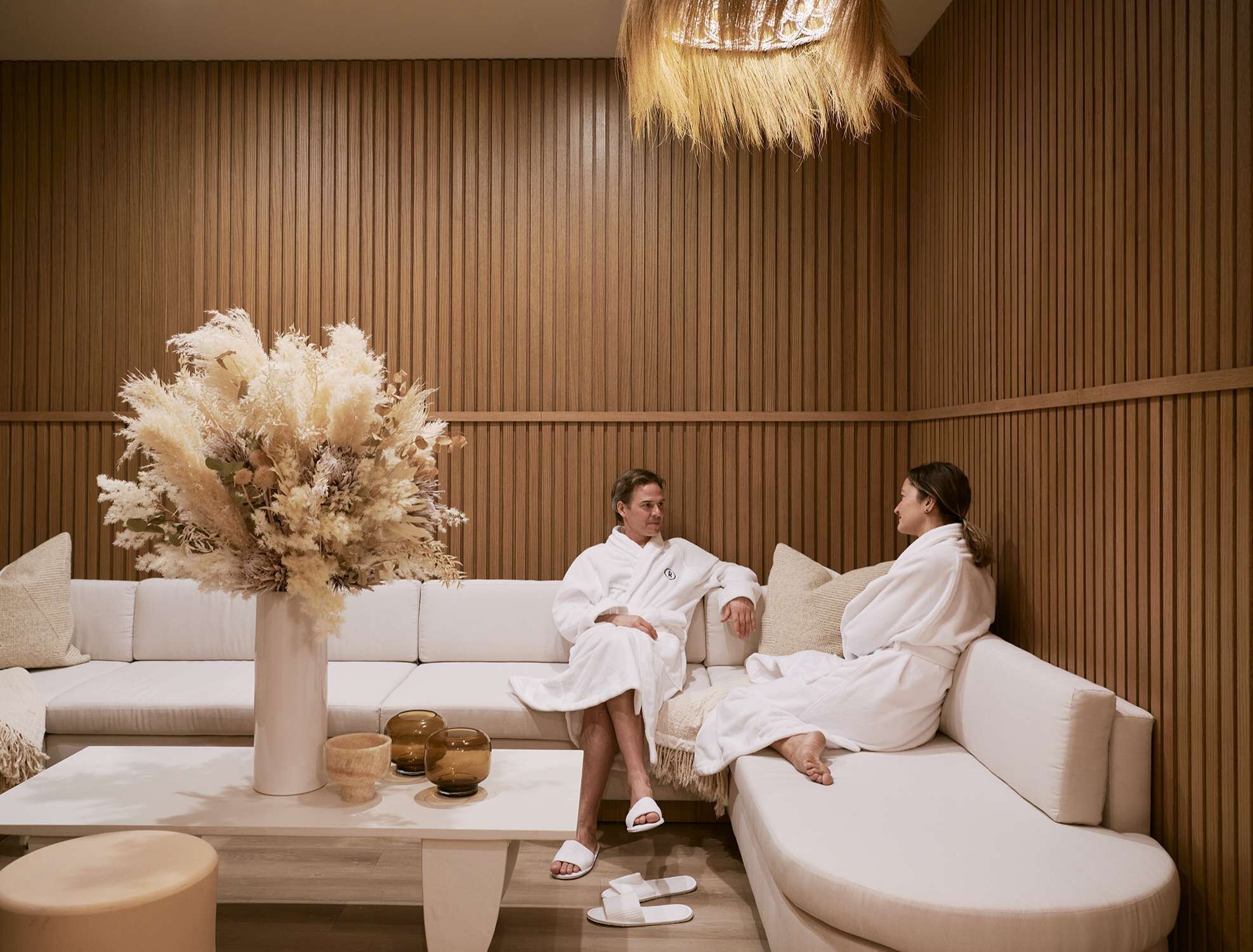 A spa room with a wooden panel wall, a white sofa, and a large vase of pampas grass.